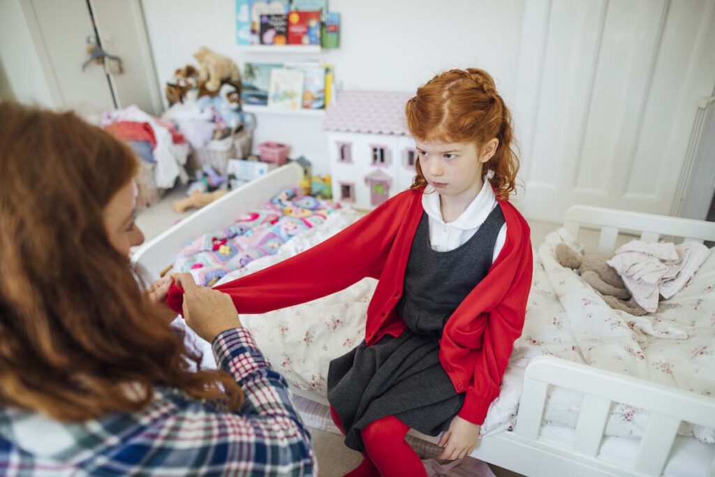 A medium shot of a mid adult, Caucasian mother trying to get her daughter to out her school cardigan on to get her ready for her first day of school. The girl is sat on the bed looking annoyed and displeased.