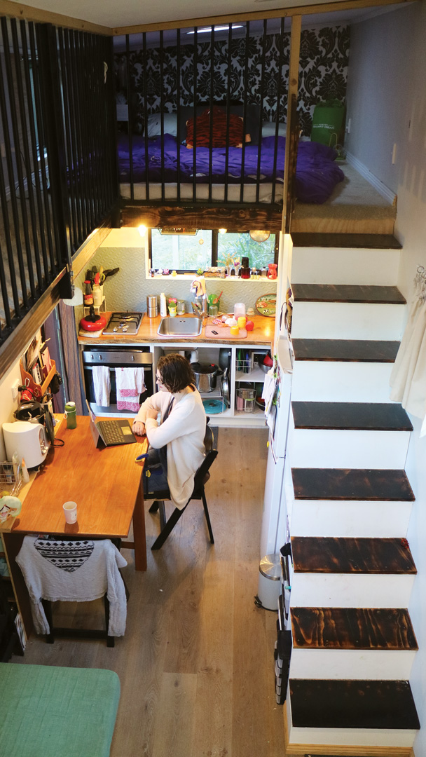 Tiny house with kids view from loft level, across to parents' bedroom, looking down at kitchen/dining, with flight of stairs on the right.
