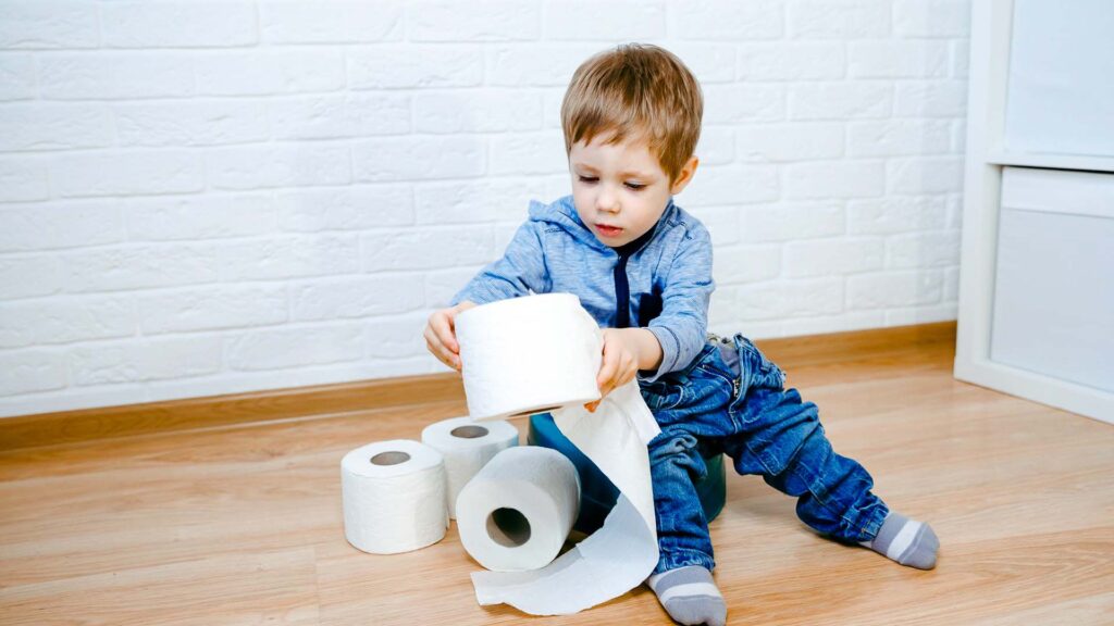 Constipated toddler sitting on a potty holding up a roll of toilet paper, with three more rolls on the ground