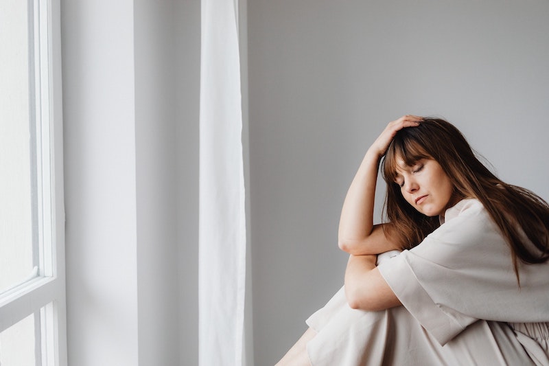 woman sitting facing a window, hugging her knees with one hand on her head. her eyes are closed and she looks upset