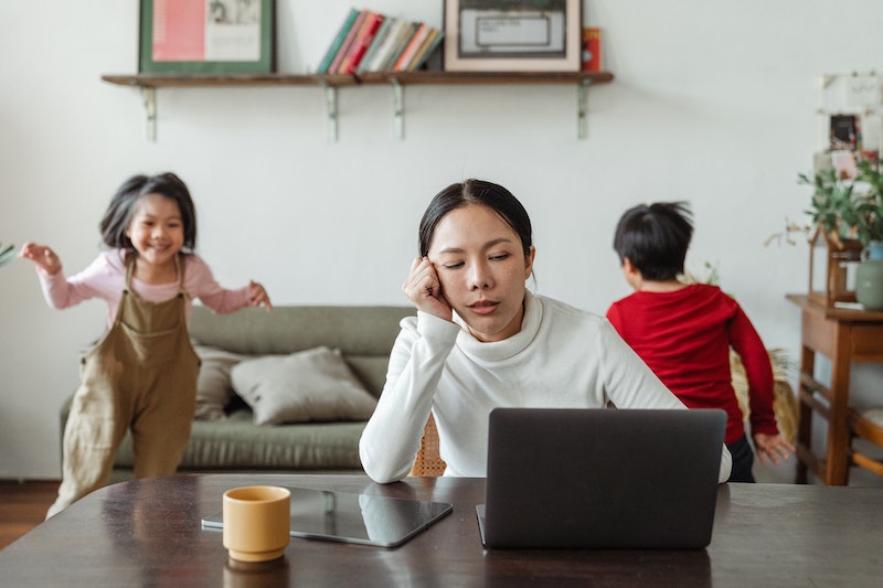 woman looking stressed sitting in front of a laptop while two children run gleefully around behind her in the living room