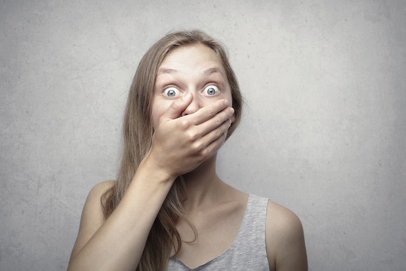 Woman standing against a wall with eyes wide open in surprise and hand covering her mouth