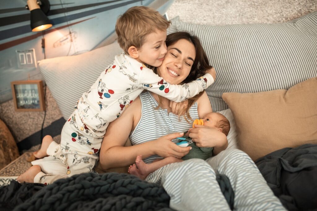 Boy hugging mother who is sitting on a couch and carrying a baby