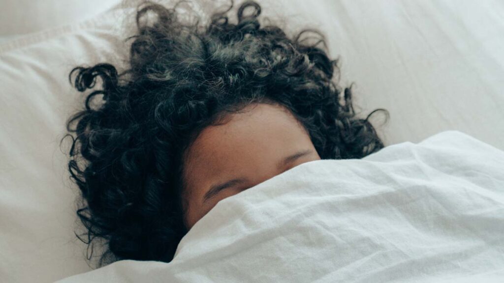 Person lying in bed with blanket covering face, only forehead is showing