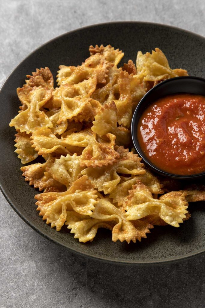 Bowl of pasta chips in a dark grey bowl on a grey slate table, with a small dipping bowl of tomato sauce
