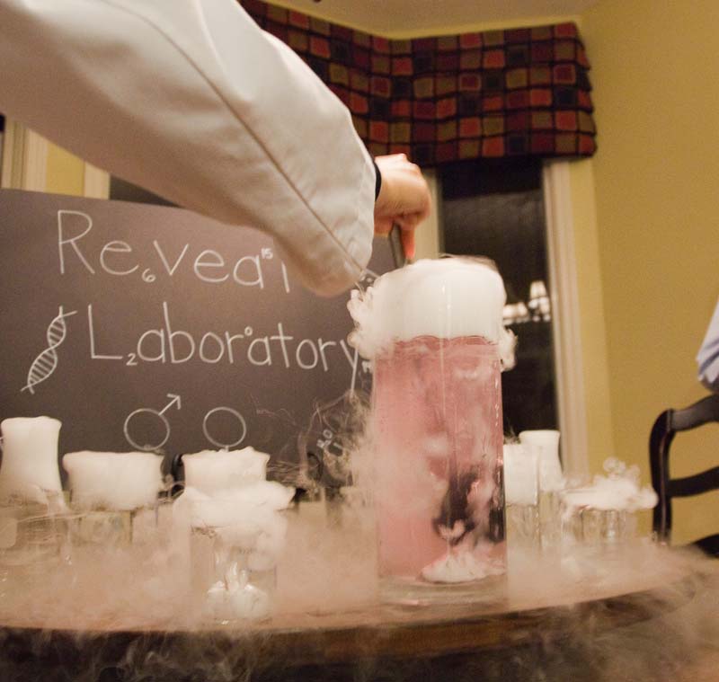 A hand stirring a tall glass pitcher filled with pink liquid and smoke bubbling over.