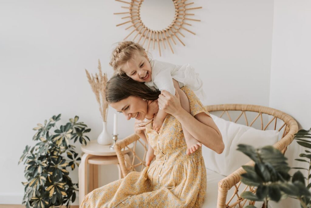 Mum is smiling and sitting on a chair and her toddler is on her back with a look of joy and exhilaration on her face.